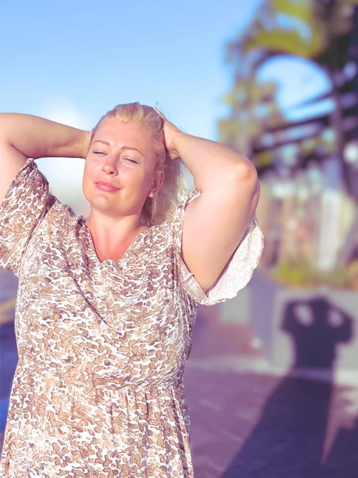 Isabell Lorenscheit steht in der Sonne, die Hände am Hinterkopf, in einem hellen Kleid mit braunem Muster. Der Hintergrund ist unscharf.